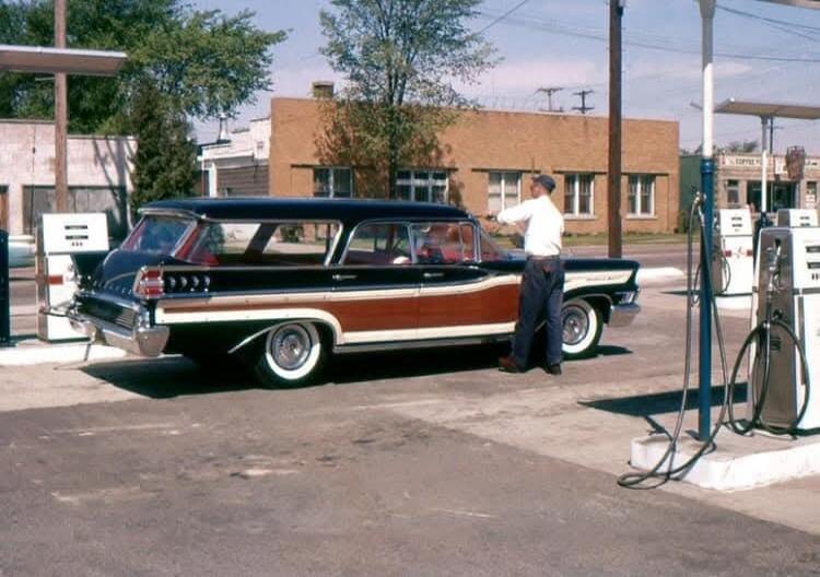 A man stands near a classic wood-paneled station wagon at a gas pump.