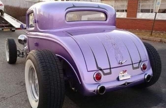 A custom purple hot rod car with large rear tires and white sidewalls is parked on asphalt. The car features a classic coupe body style with custom pinstriping on the trunk. A car trailer is visible in the background.