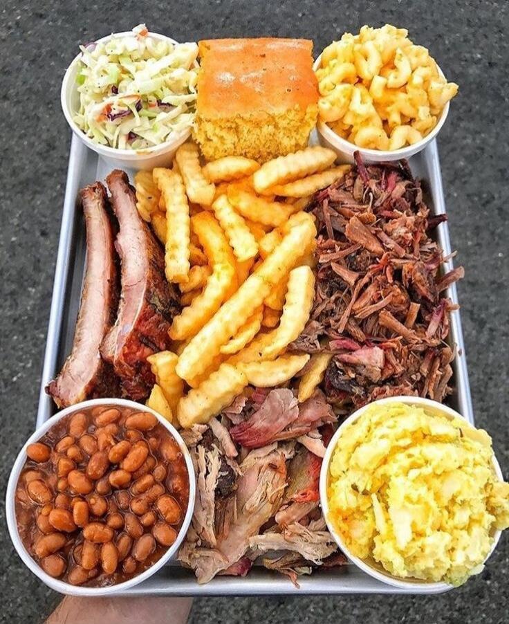 A large metal tray filled with a variety of barbecue dishes and sides. It includes several racks of ribs, a generous portion of pulled pork or brisket, and a pile of golden french fries. Accompanying these are small bowls of coleslaw, macaroni and cheese, baked beans, and mashed potatoes. A square piece of cornbread is also visible.