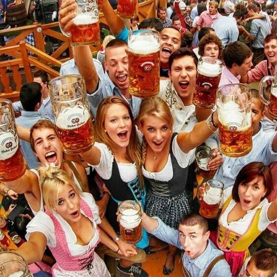 A large group of people, mostly young adults, are celebrating and cheering, holding up large mugs of beer. Many are wearing traditional Bavarian clothing like dirndls and lederhosen. The text 'Hacker-Pschorr' is visible on some beer mugs.