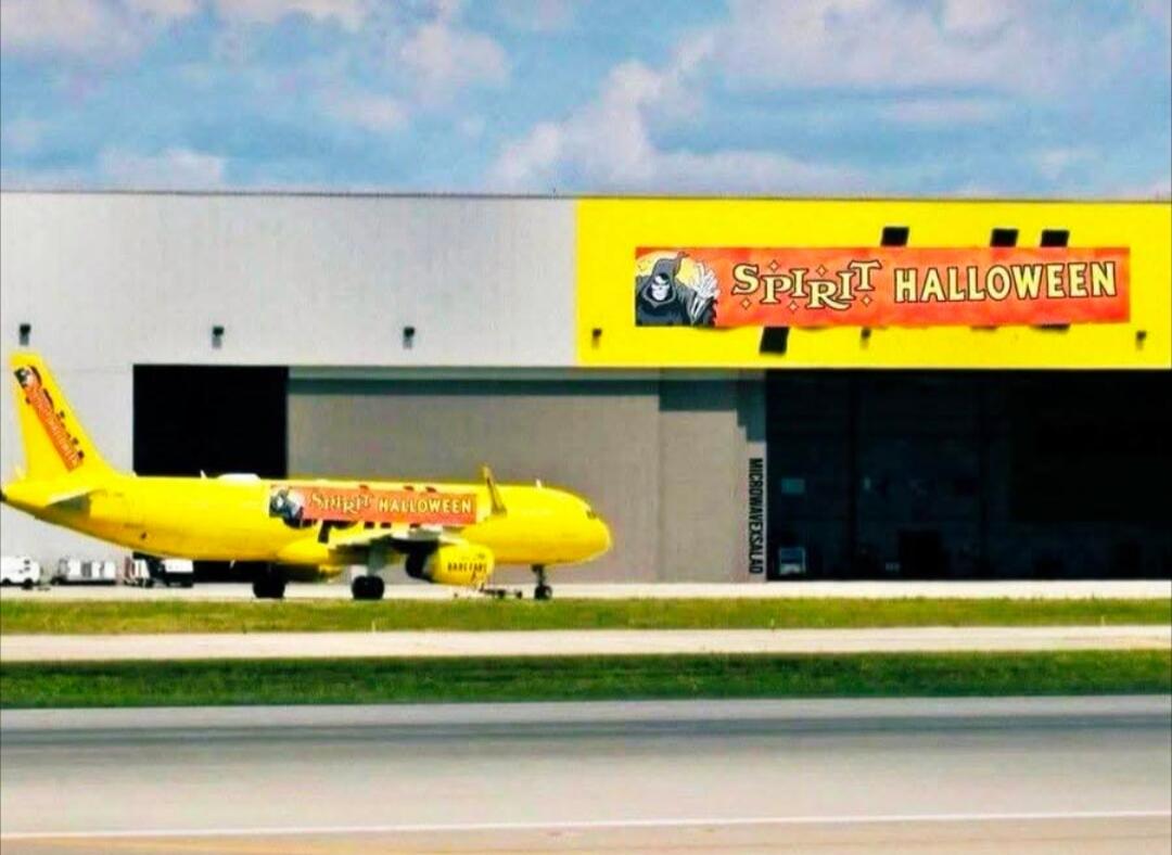 A yellow airplane resembling a Spirit Airlines plane is shown on a runway next to a building. Both the plane and a large banner on the building feature the 'Spirit Halloween' logo and text, creating a humorous visual pun.