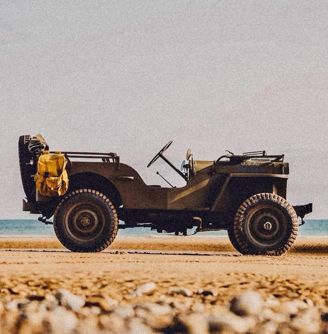 An old military-style jeep sits on a sandy beach with the ocean in the background.