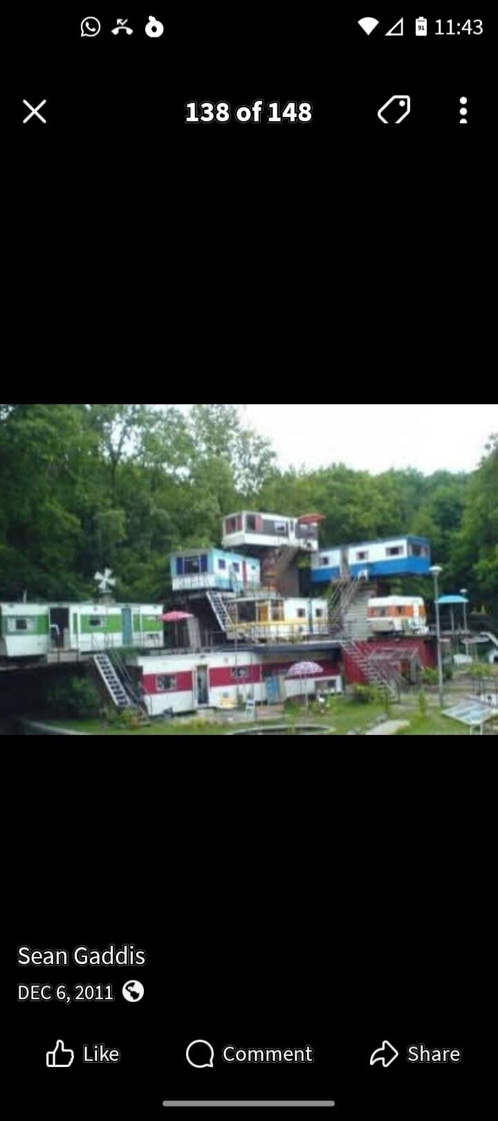 A unique and unconventional multi-level structure built from various colorful trailers or caravans stacked and connected on a green hillside. The image also contains text: Sean Gaddis, DEC 6, 2011, Like, Comment, Share, 138 of 148, 11:43.