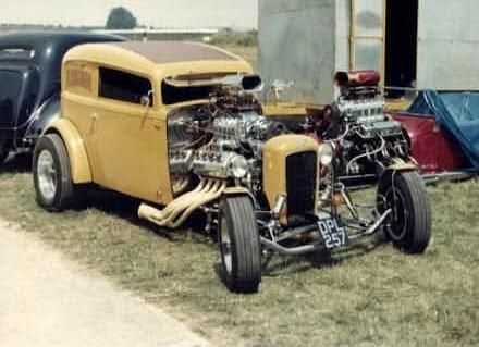 A vintage yellow hot rod with a large exposed engine and prominent exhaust pipes is parked on a grassy field. Another custom car is partially visible behind it. The license plate on the front of the yellow car reads DPL 257.