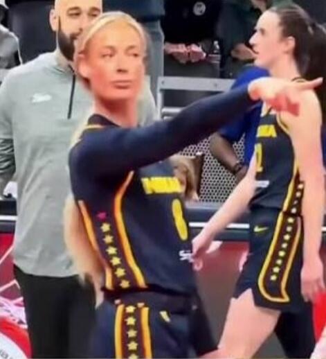 A blonde female basketball player in a dark blue and yellow uniform is looking forward, extending her arm and pointing. Another player is visible behind her on the court.