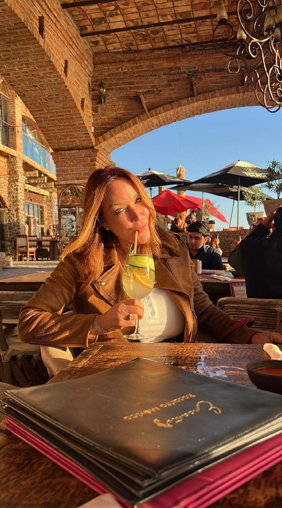 A woman is sitting at an outdoor restaurant, holding a drink with a straw. The menu on the table says 'Rosarito México'.