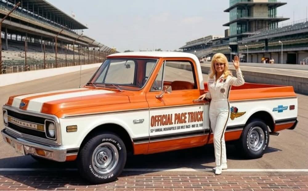 OFFICIAL PACE TRUCK 53rd ANNUAL INDIANAPOLIS 500 MILE RACE - MAY 30, 1969 CHEVROLET C-10 GOODRICH Radial T/A. A woman in a white racing suit stands next to a vintage orange and white Chevrolet C-10 pickup truck, which served as the official pace truck for the Indianapolis 500 race. The scene is set on a race track with grandstands and a control tow
