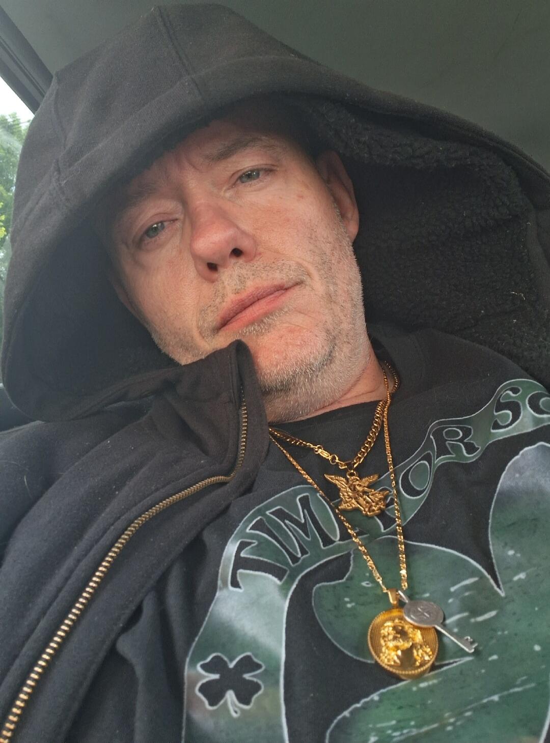 A man wearing a black hoodie and a black t-shirt with a green shamrock design and the word 'TIMOR'. He has a short beard and is wearing multiple gold chains with pendants, including an angel and a coin with a key.