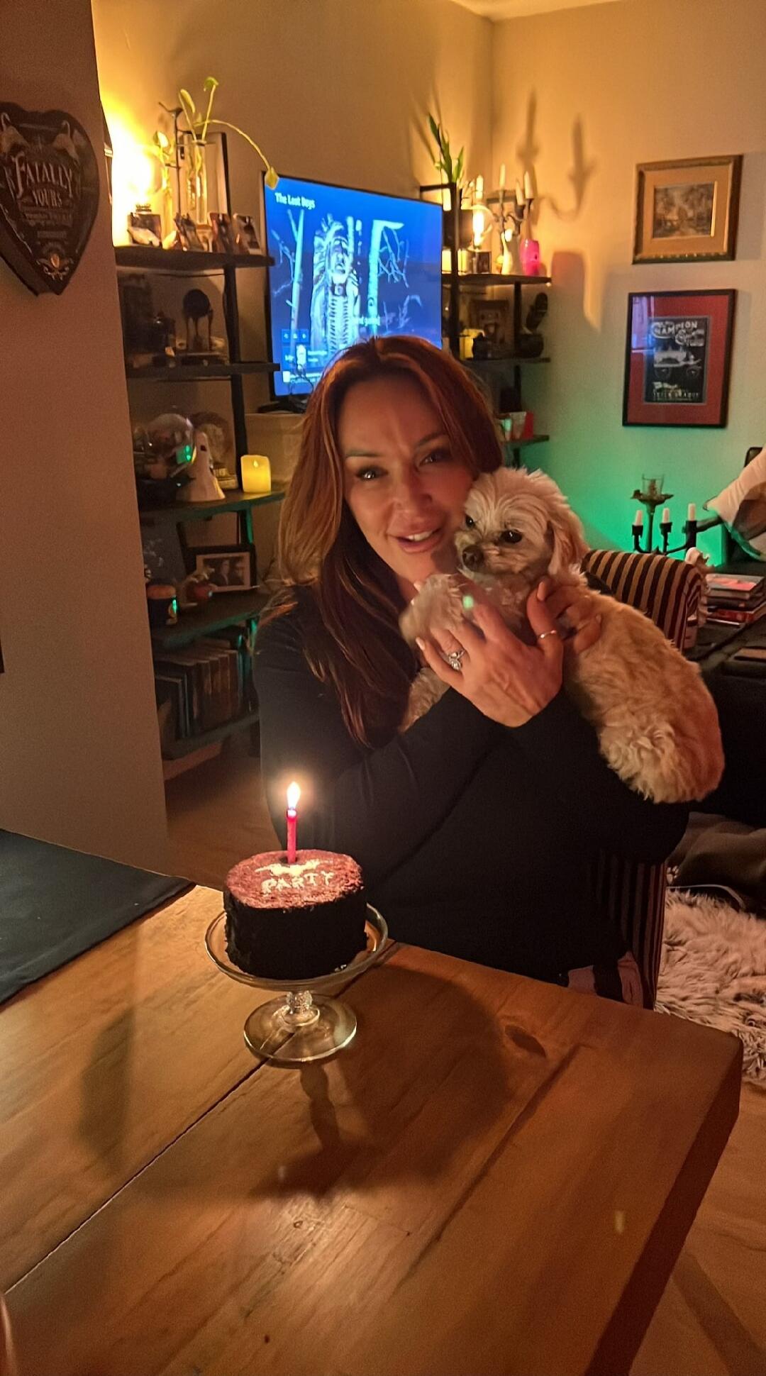 FATALLY YOURS, The Lost Boys, PARTY. A woman is smiling and holding a small dog, with a lit candle on a small cake in front of her, suggesting a celebration.