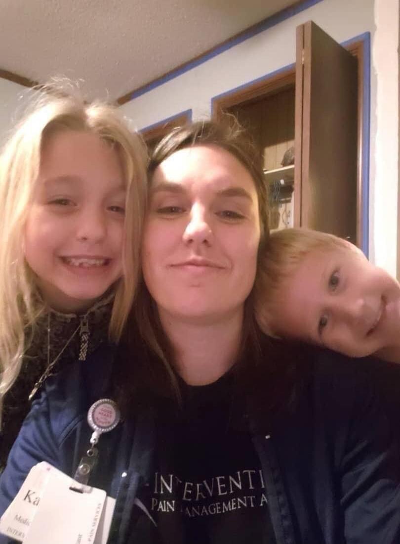 Three people posing for a selfie indoors. A woman is in the center with a slight smile, flanked by a smiling girl on the left and a boy leaning his head on her shoulder on the right. The woman is wearing a dark top with a jacket; the girl on the left has long blonde hair and is wearing a badge or lanyard; the boy on the right has light hair and is 