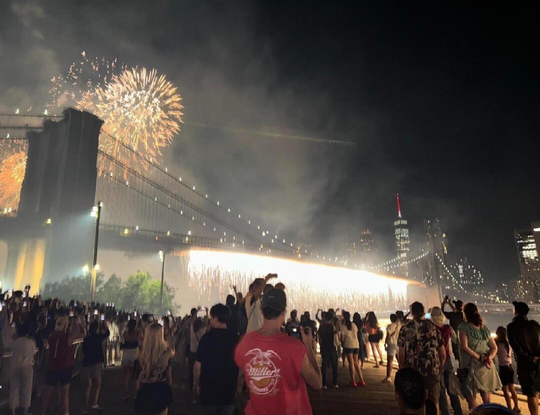 A large crowd of people gathered at night to watch a spectacular fireworks display over the Brooklyn Bridge, with the One World Trade Center visible in the background. Fireworks are exploding in the sky and cascading from the bridge structure. A person in the foreground wears a shirt with 'Miller's' text.