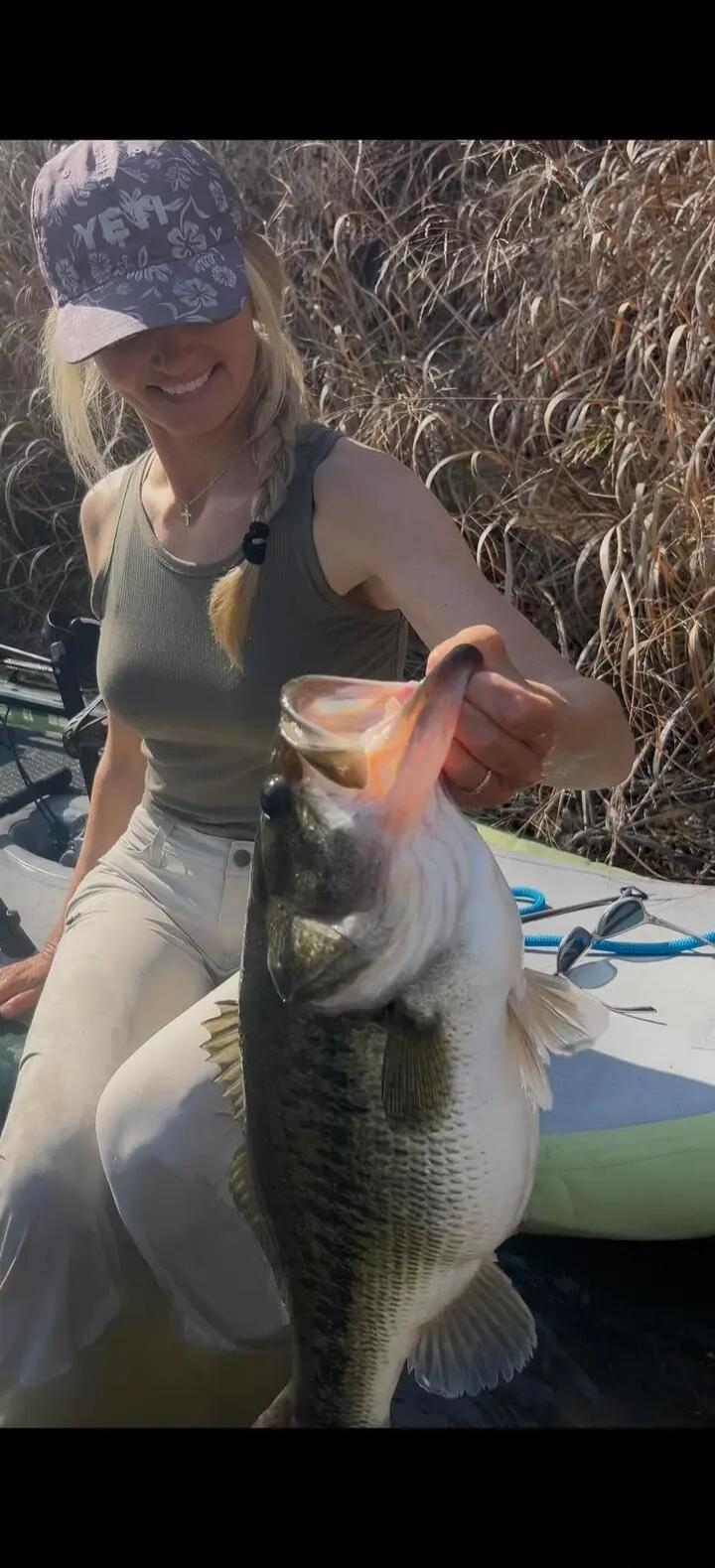 A woman wearing a YETI cap and a tank top is holding a large bass fish. She is smiling and appears to be on a boat or kayak, with dry reeds in the background.