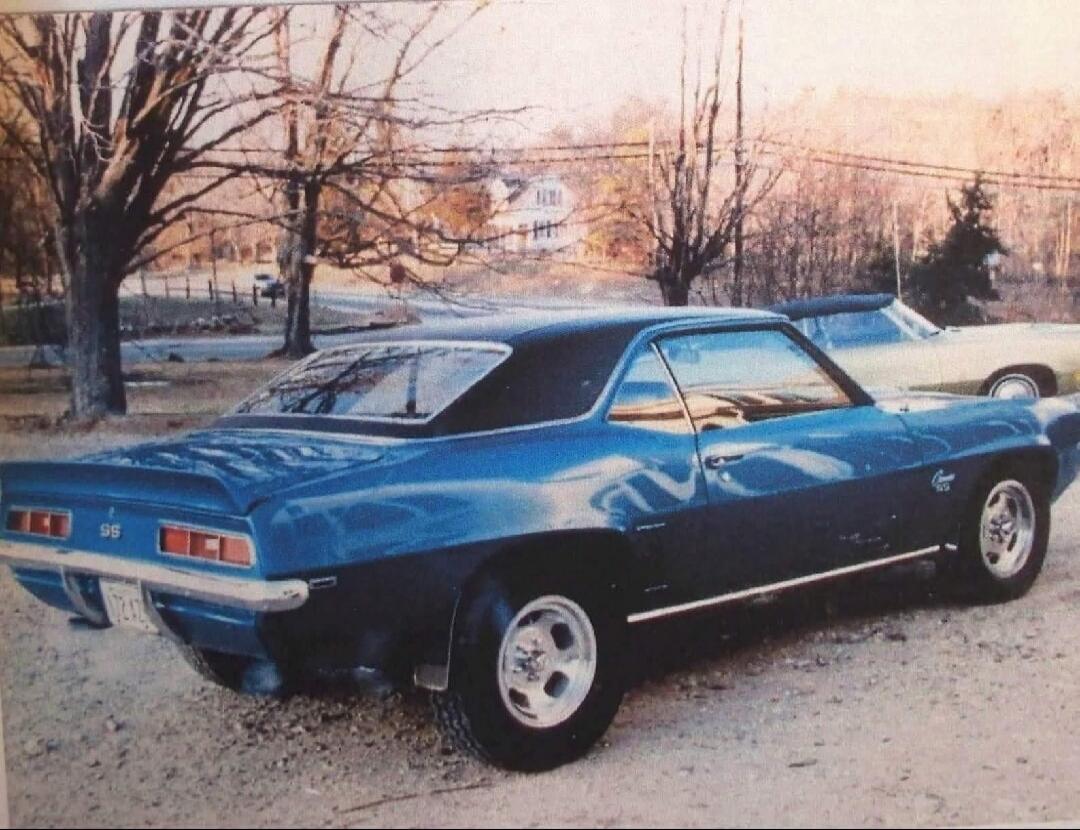 A blue classic Chevrolet Camaro SS parked outdoors on a gravel surface. The car has a black vinyl roof and custom wheels. Another car is partially visible in the background.