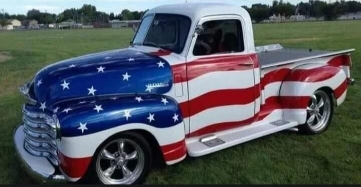 The image shows a classic pickup truck painted in the style of the American flag. The front of the truck, including the hood and fenders, is painted blue with white stars, resembling the union of the flag. The sides of the truck feature red and white stripes, mirroring the stripes of the flag. The truck appears to be a vintage model, possibly from 