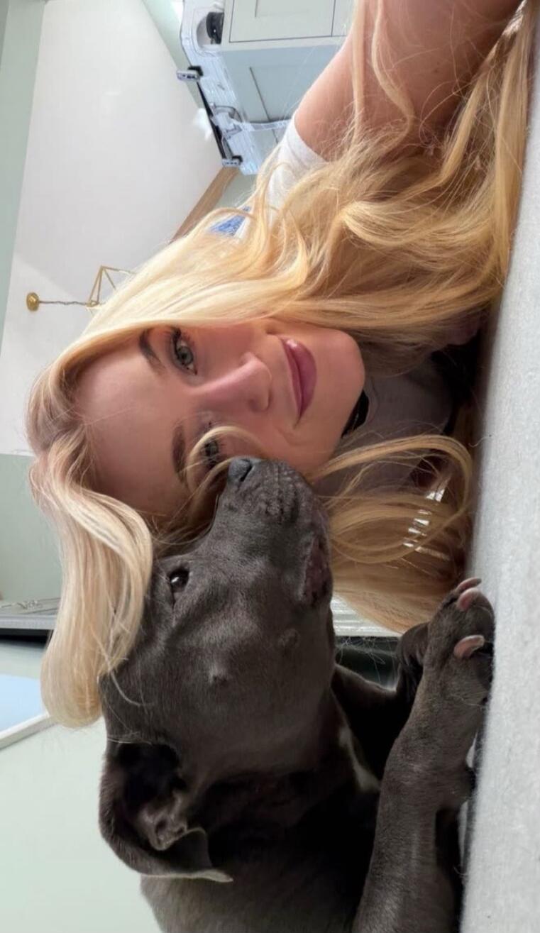 A blonde woman lying on a surface next to a gray dog, both looking at each other with the dog resting its head close to the woman's face. They appear close and affectionate.