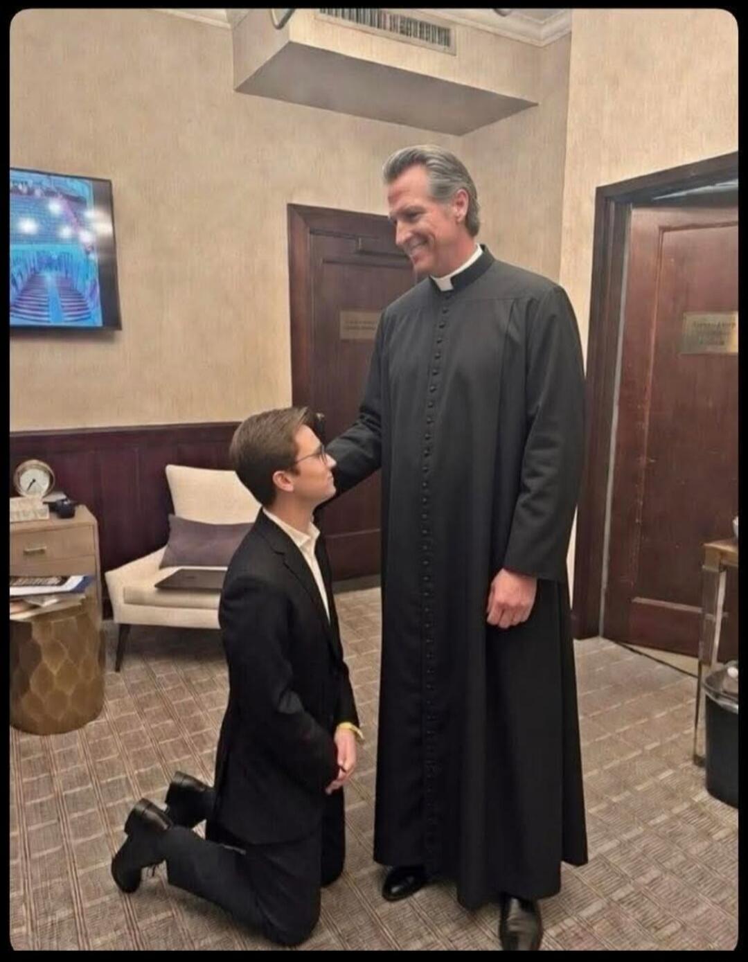 A young boy kneels in front of a tall clergyman in a long black robe, who is smiling and shaking his hand in a hallway or lounge area.