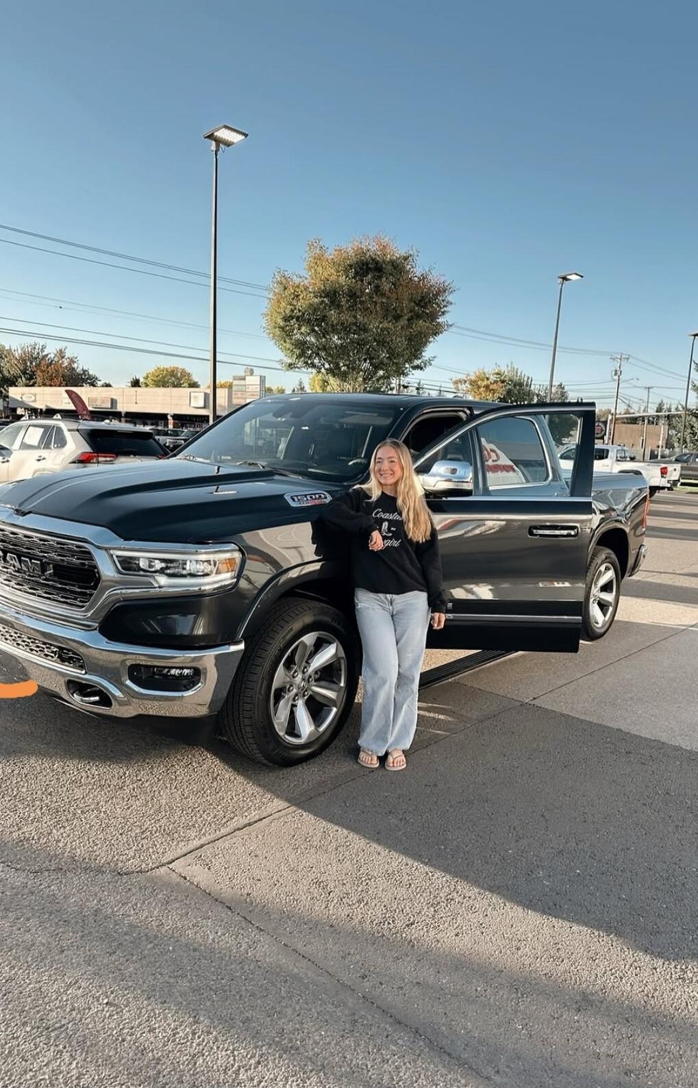 A young woman with long blonde hair is smiling and leaning on a black RAM 1500 pickup truck in a parking lot. The truck's driver-side door is open. She is wearing a black sweatshirt that says 'Coaster Girl' and light-colored jeans. The sky is clear blue.