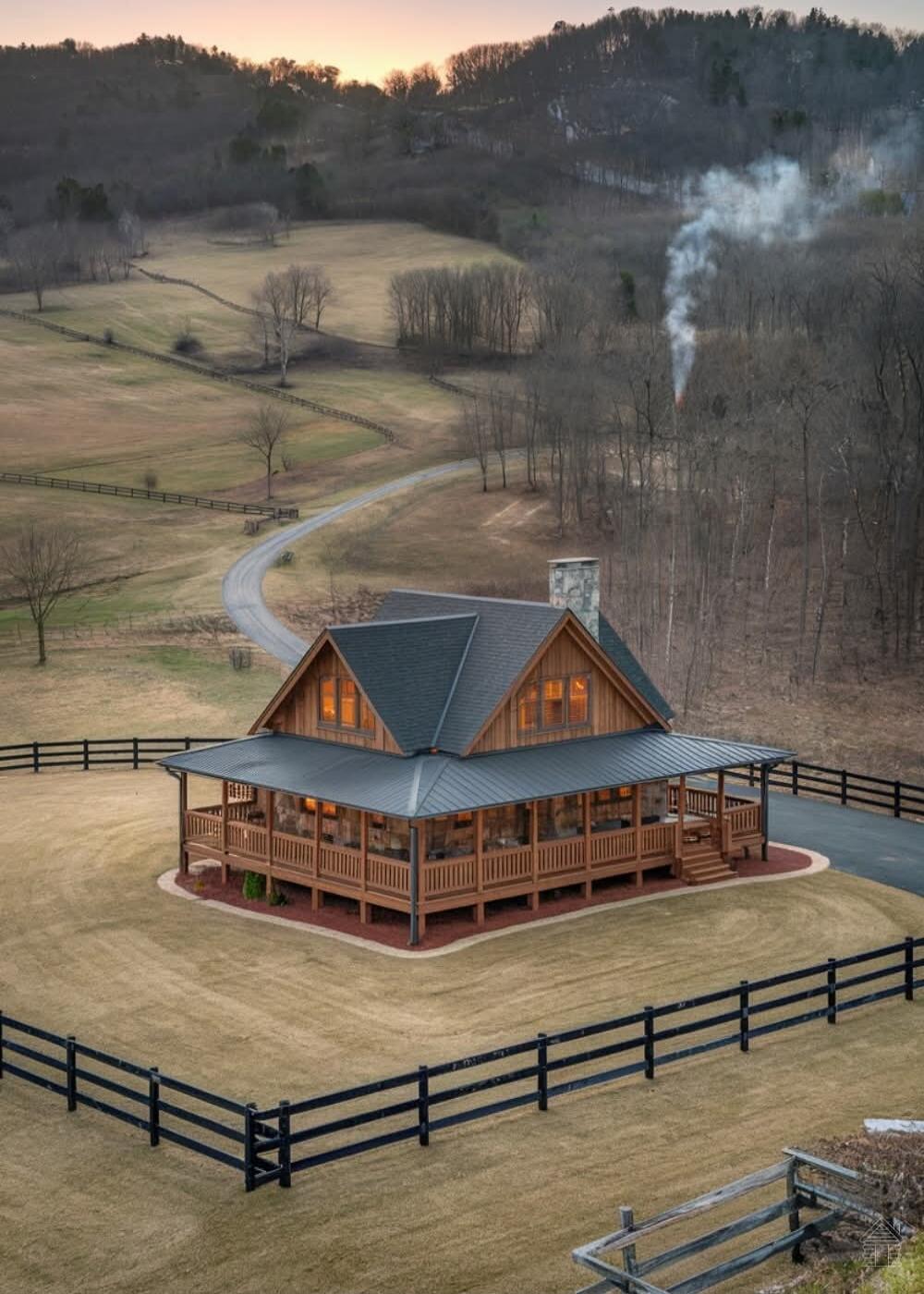 A wooden cabin with a wraparound porch sits on a circular stone foundation inside a fenced yard. Smoke rises from the chimney on a rural property surrounded by rolling hills, leafless trees, and a winding road. The scene is bathed in warm light from a setting or rising sun.