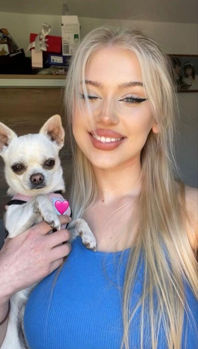 A smiling blonde woman wearing a blue top is holding a small white dog with a pink heart emoji on its collar.