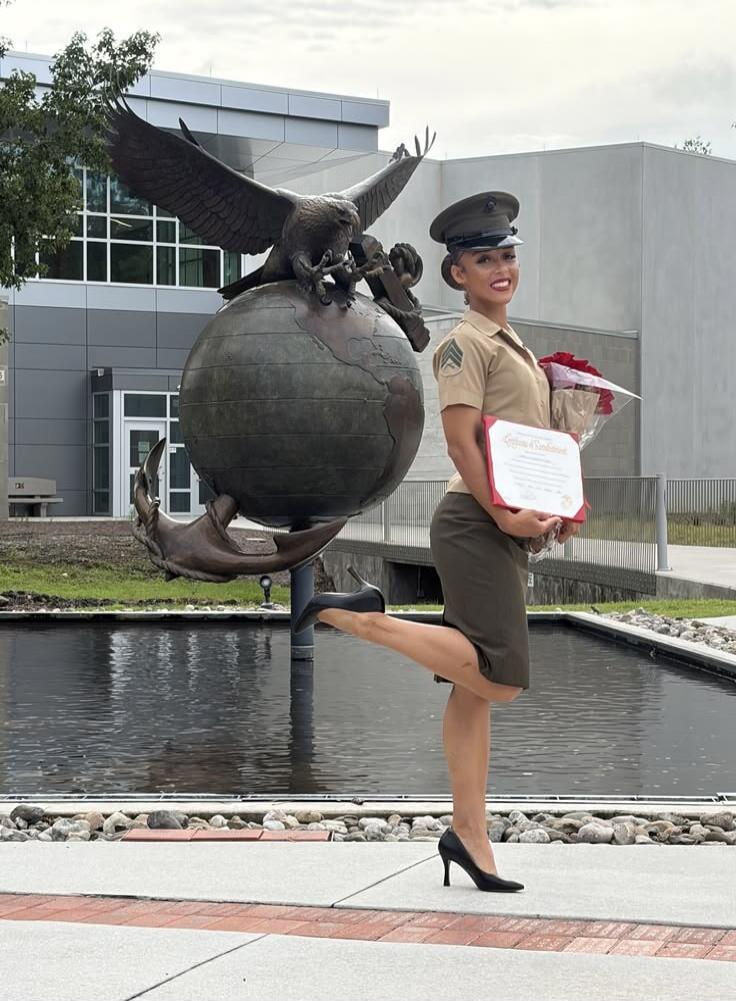 A woman in a US Marine Corps uniform stands next to a large statue of an eagle, globe, and anchor. She is holding a certificate and a bouquet of red roses, posing with one leg lifted. The certificate appears to be a 'Certificate of Commendation'.