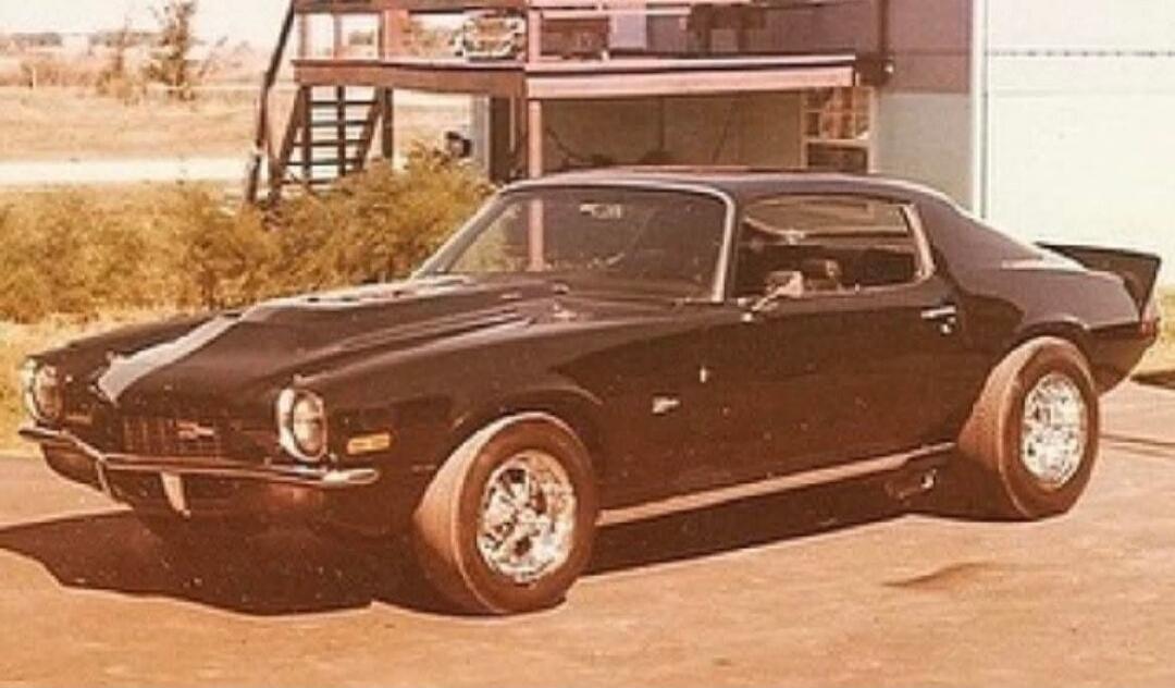 A black vintage muscle car parked outside, with wide rear tires and chrome wheels, on a sunny day near a two-story building.