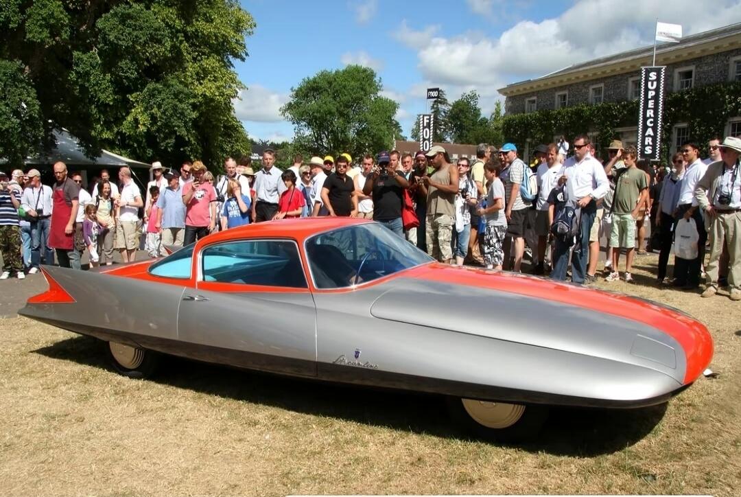 A silver and red futuristic car on display outdoors, surrounded by a large crowd of people.
