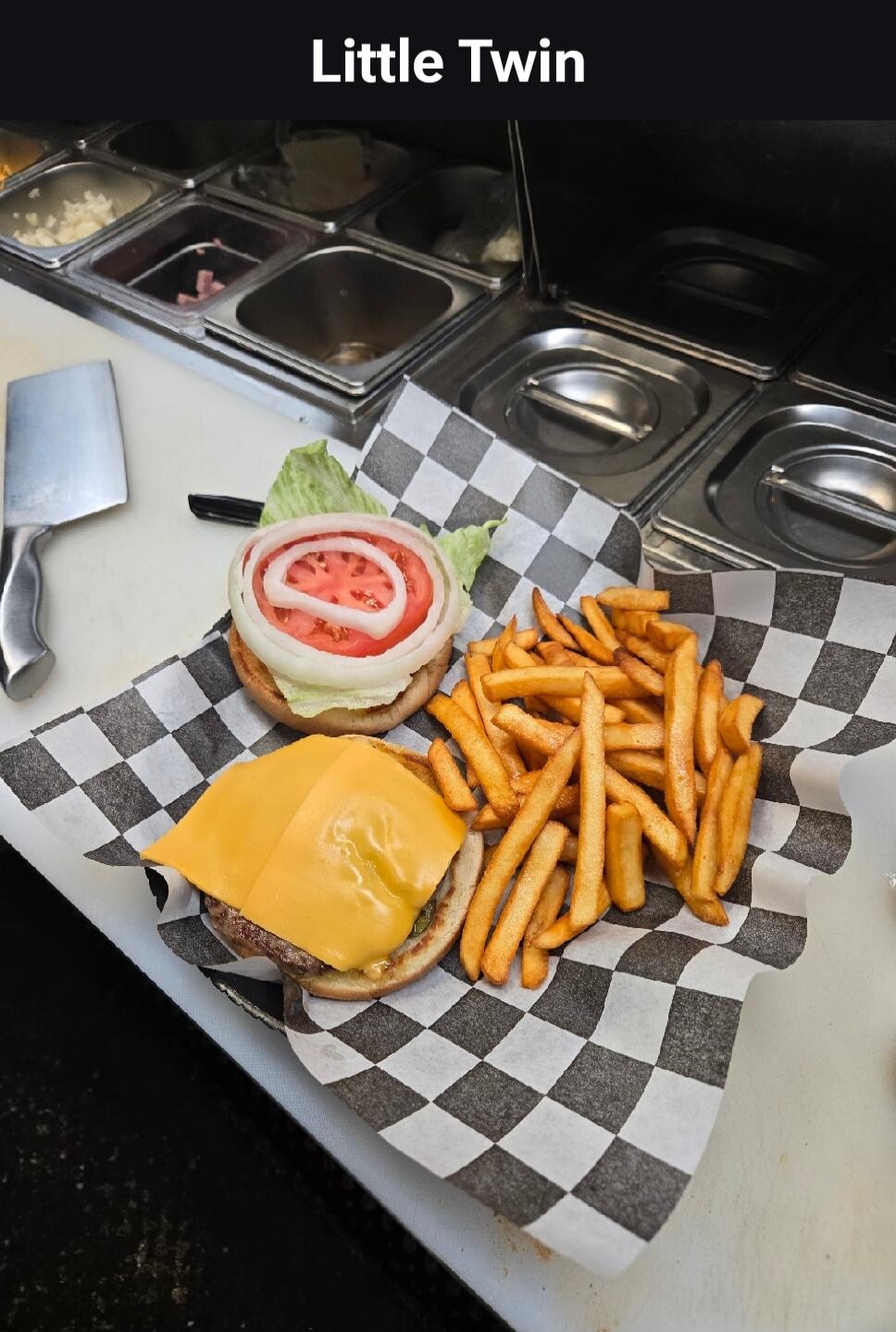 Little Twin. An open burger with cheese, lettuce, tomato, and onion, served with french fries on checkered paper. A cleaver and stainless steel containers are visible in the background.