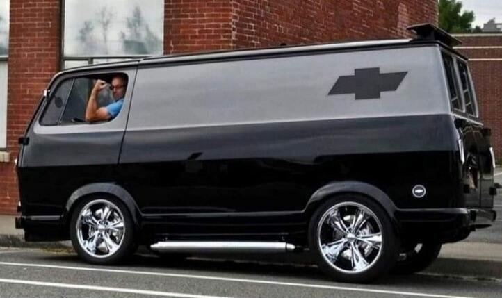 A man in a customized black and grey Chevrolet van is flexing his bicep out the driver's window. The van features large chrome wheels and side exhaust pipes. A black Chevrolet logo is visible on the side.