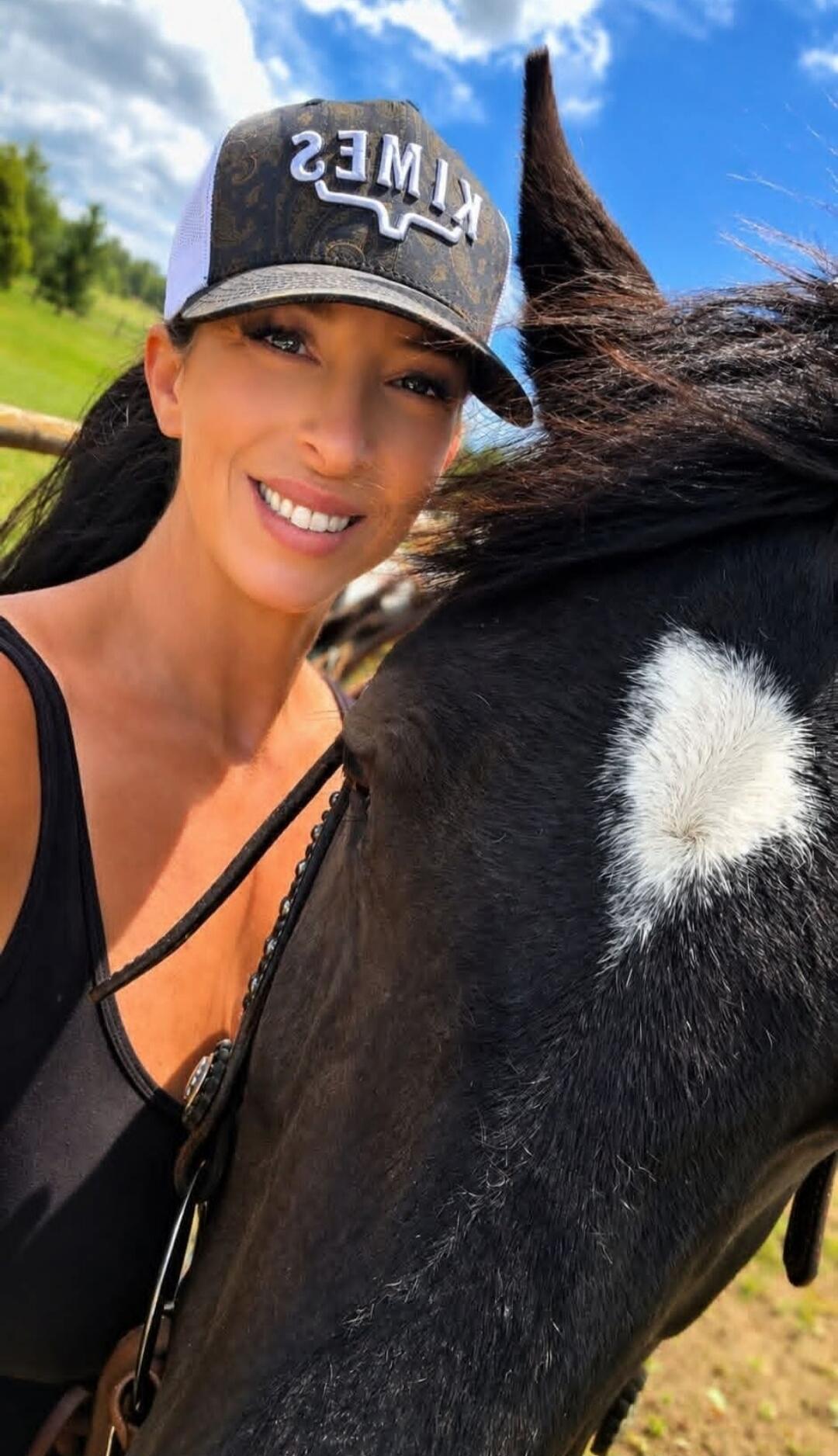 A woman in a black tank top and a baseball cap with 'KIMES' written on it, smiling next to a black horse with a white patch on its forehead.