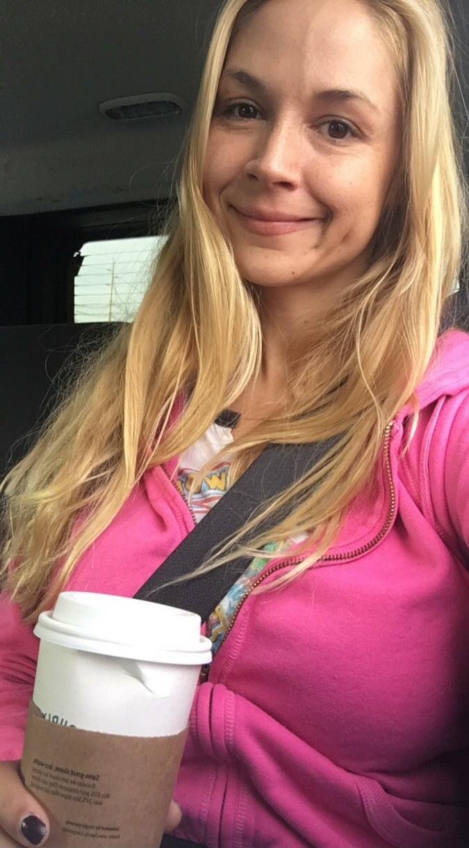 A woman in a car, wearing a pink hoodie and a seatbelt, holding a coffee cup. The text on the cup is mostly illegible, but 'TIDIA' is visible upside down.