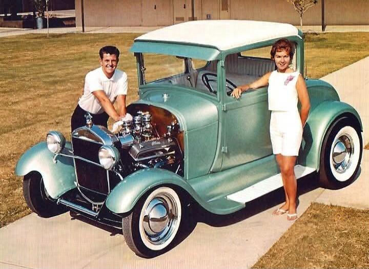 A vintage photograph showing a smiling man and woman posing with a classic custom hot rod car. The man is leaning over the engine bay, which is open, revealing a modified engine. The woman stands beside the car door. The car is a light green or teal color with a white roof and whitewall tires. They appear to be proud of the vehicle.