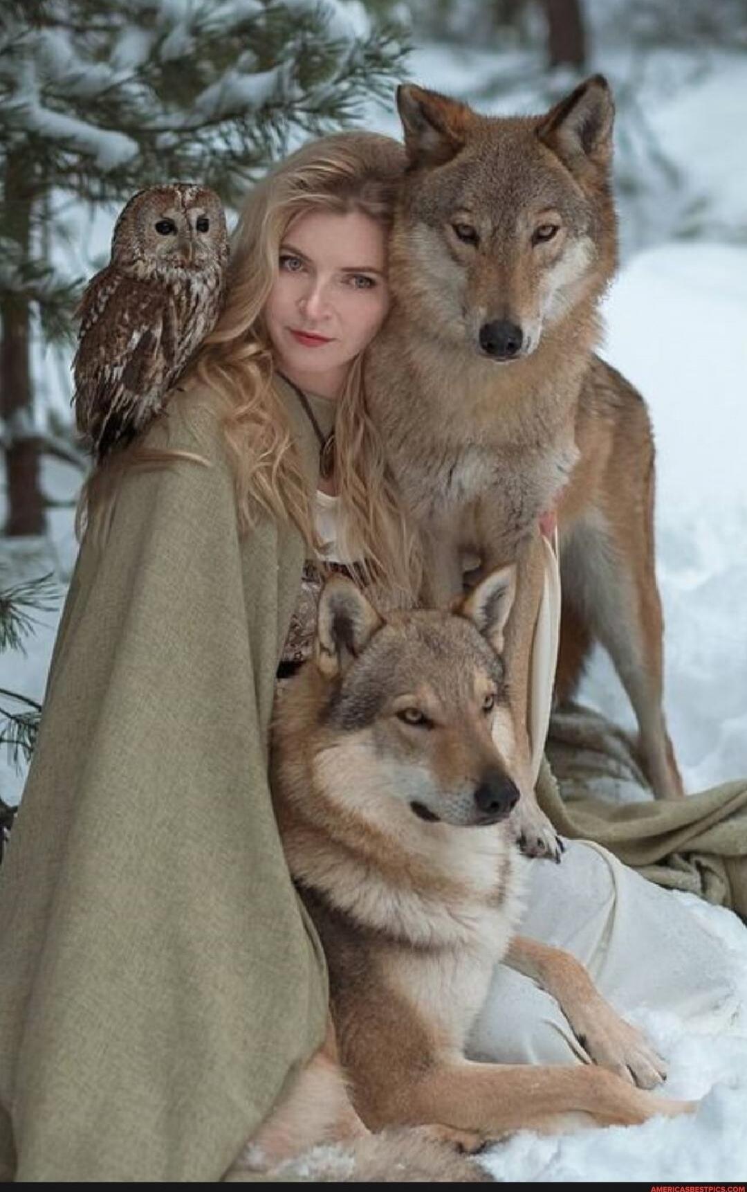 A woman sits in the snow with two wolves and an owl perched on her shoulder.