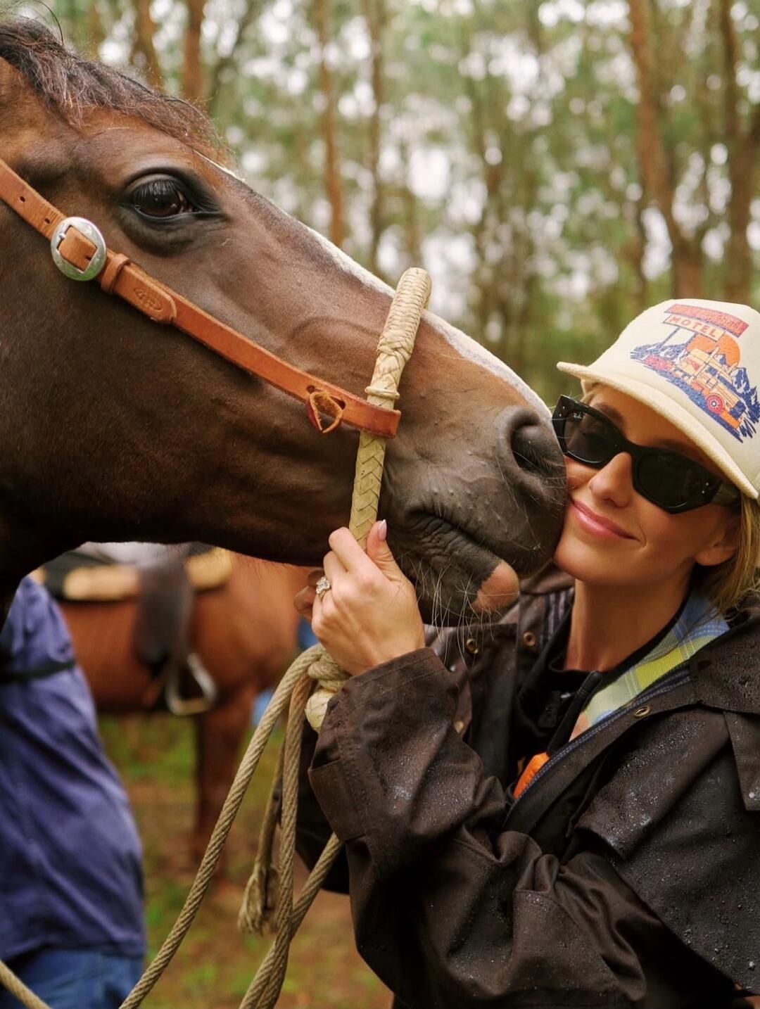 A woman in sunglasses and a hat kissing a horse on the nose outdoors.