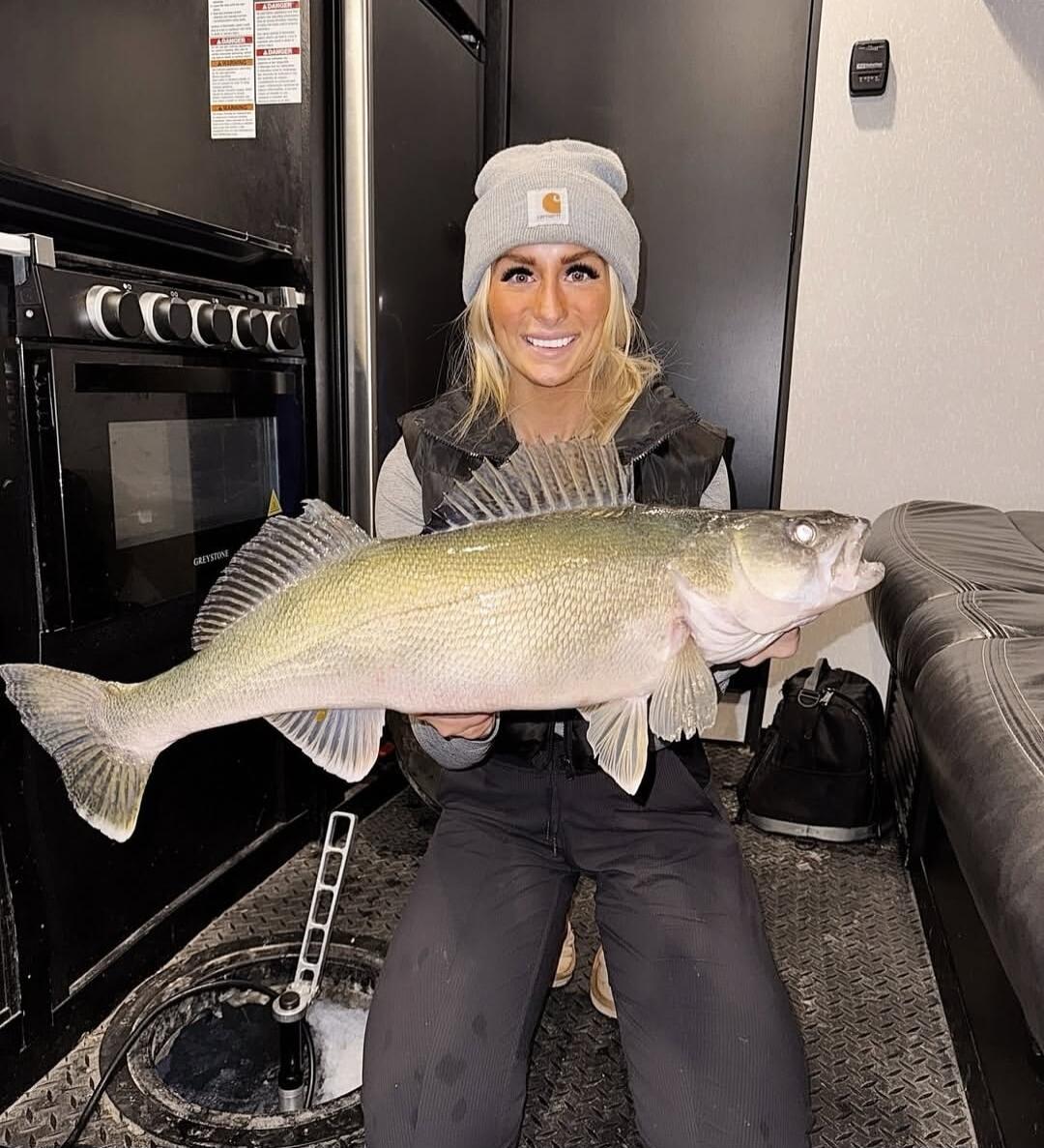 A woman in winter attire, including a grey beanie and a dark vest, kneels inside what appears to be an ice fishing shelter, proudly holding a large walleye. An ice fishing hole with equipment is visible on the floor, and a stove/oven unit with the brand 'GREYSTONE' is in the background. She is smiling at the camera. Some warning labels, likely in E