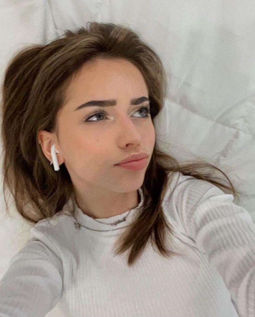 A young woman is lying down on a white surface, looking to the side. She is wearing a white ribbed long-sleeve top and a white earbud in her left ear.