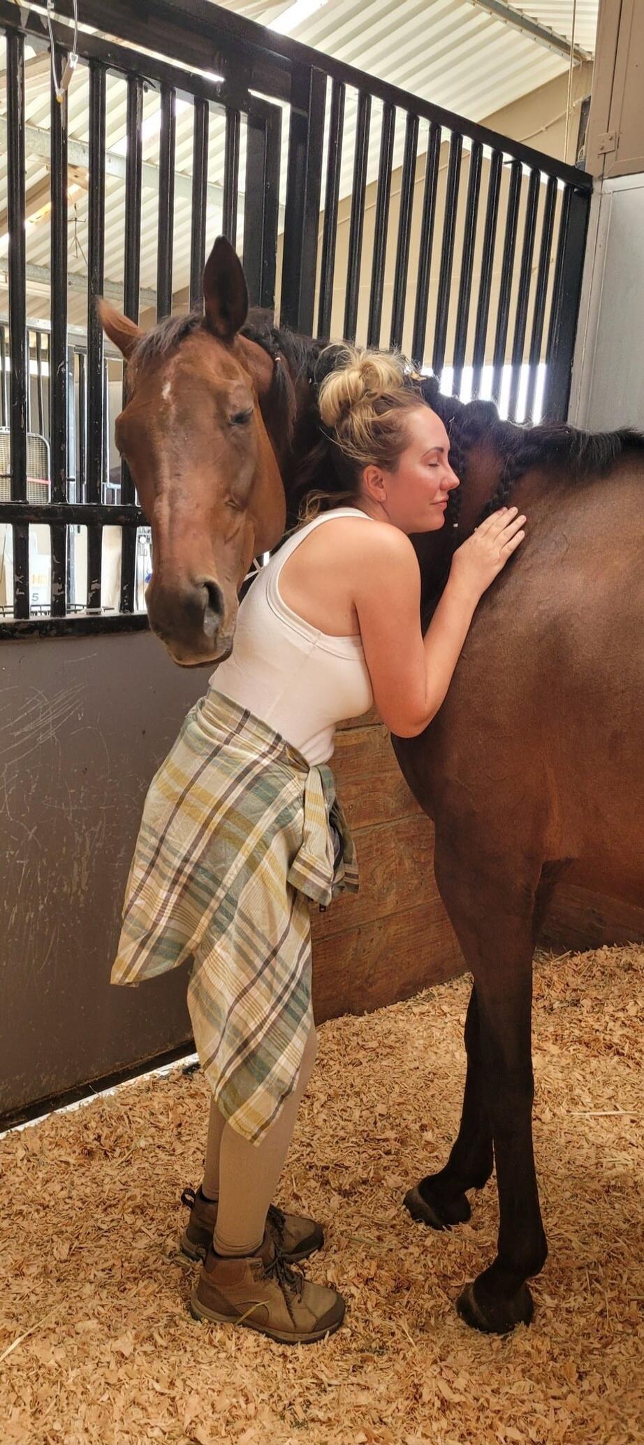 A woman in a stable is affectionately hugging a brown horse. The horse is calm and leans into her embrace. The stable floor is covered with wood shavings.