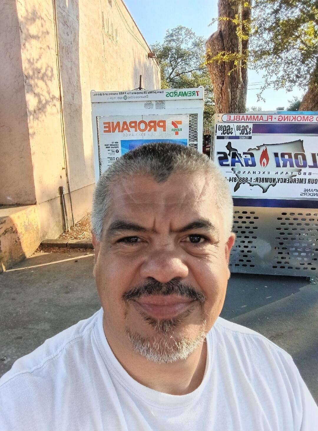 A man takes a selfie in front of two vending machines. The text on the machines, which appears reversed, includes 'PROPANE EXCHANGE', 'REWARDS', 'SMOKING & FLAMMABLE', 'LORI GAS', 'BBQ TANK EXCHANGE', and an 'EMERGENCY NUMBER'.