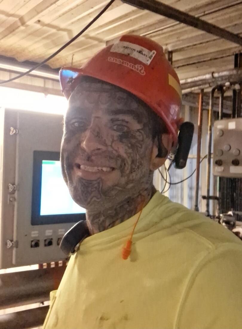 A man with a dirty face, covered in black smudges, wearing a hard hat and a yellow shirt, smiling in an industrial setting. Text on hard hat: FRANK POTTER Simmons.