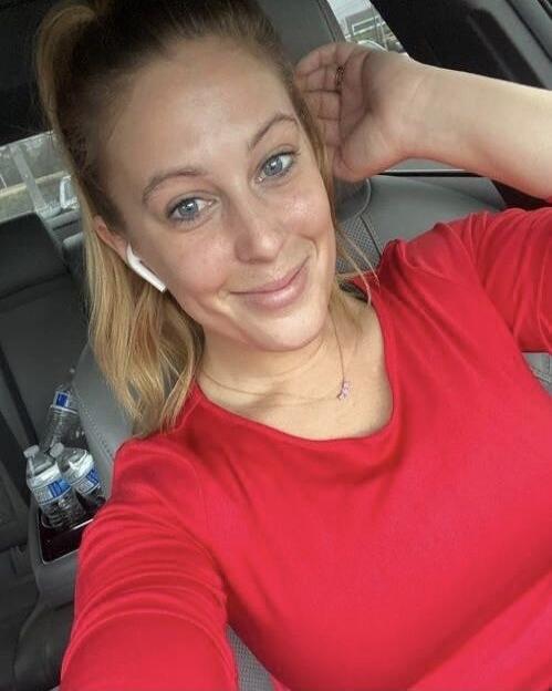 A woman with blonde hair and blue eyes, wearing a red long-sleeved top and a white earbud, smiles at the camera. She is in what appears to be a car, with water bottles visible in the background.