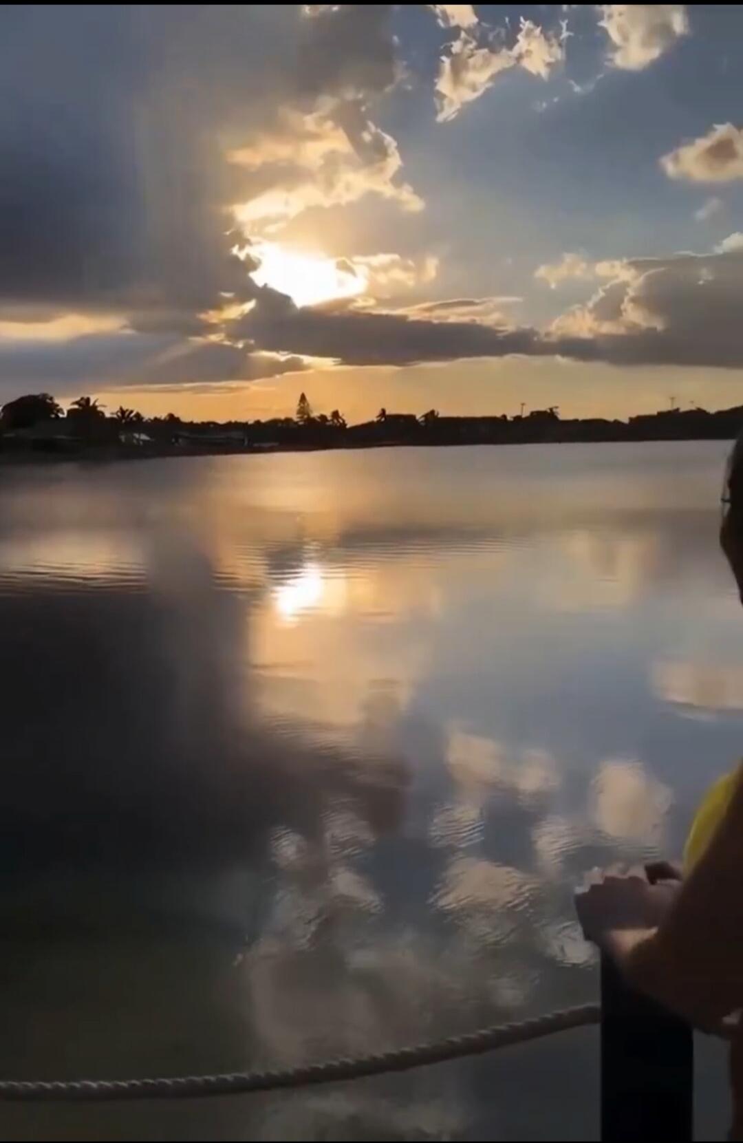 The sun is setting over the water, casting a beautiful reflection on the clouds and the lake. The sky is filled with dramatic clouds, some dark and stormy, others illuminated by the golden rays of the setting sun. A silhouette of a person can be seen on the right, looking out at the serene landscape.