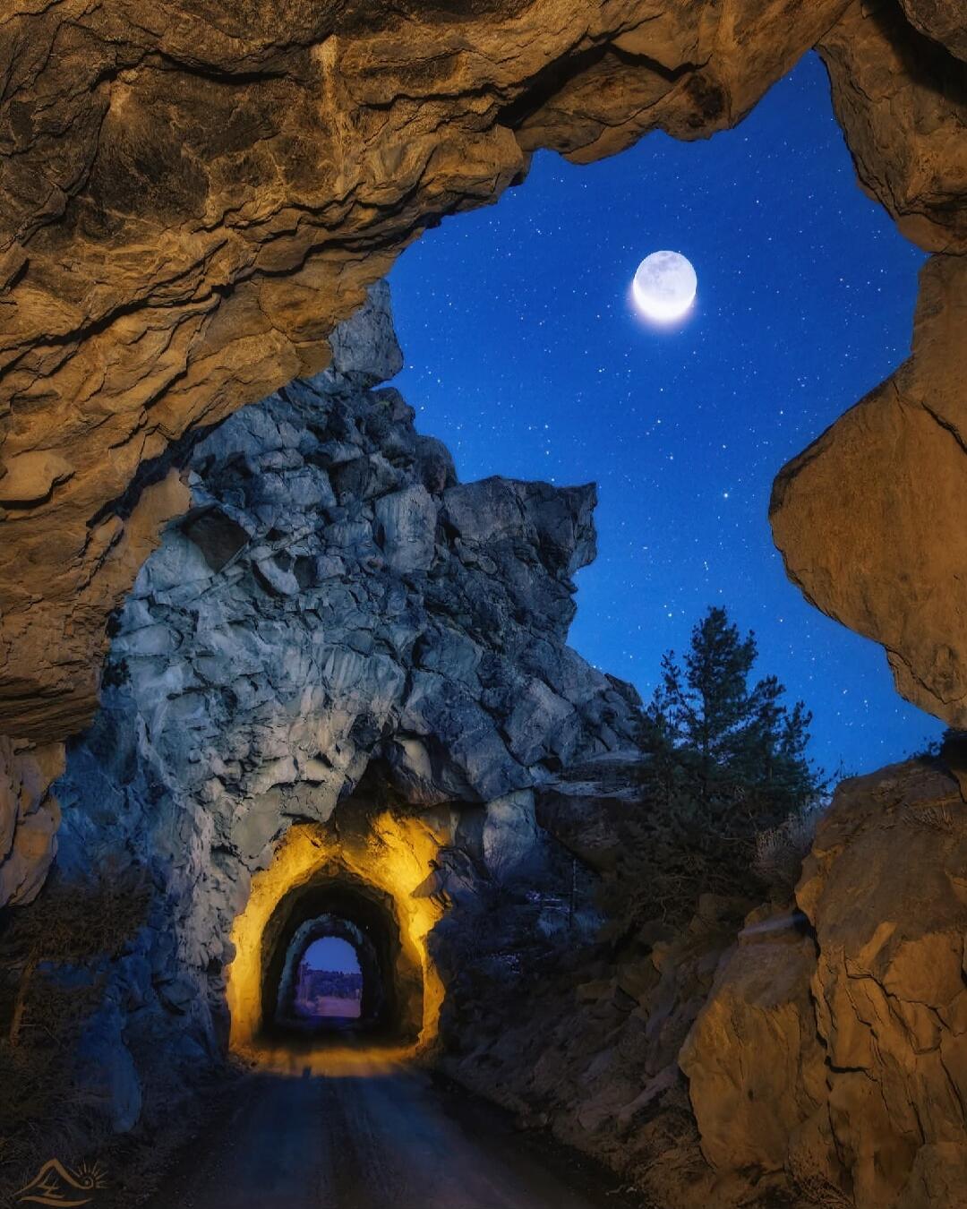 A view through a rocky archway at night, with a tunnel entrance illuminated by warm light and a full moon shining in the starry sky.
