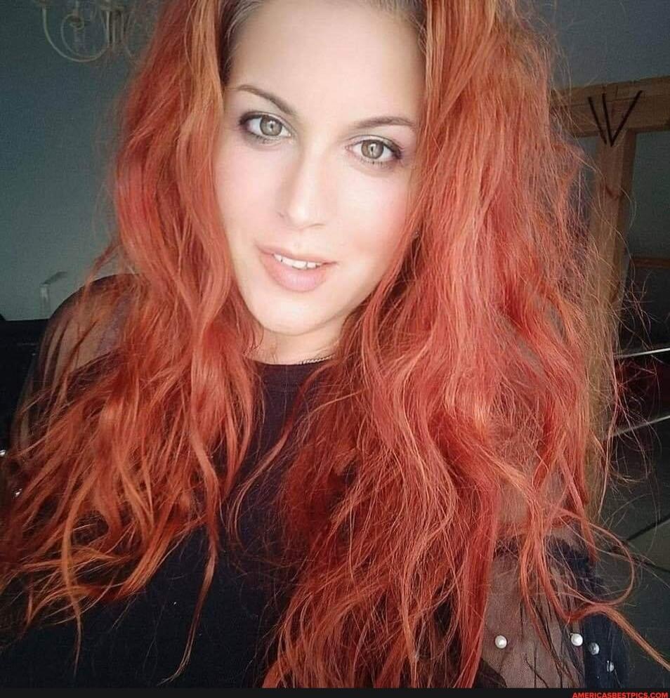 A woman with long wavy red hair wearing a black top, looking at the camera.