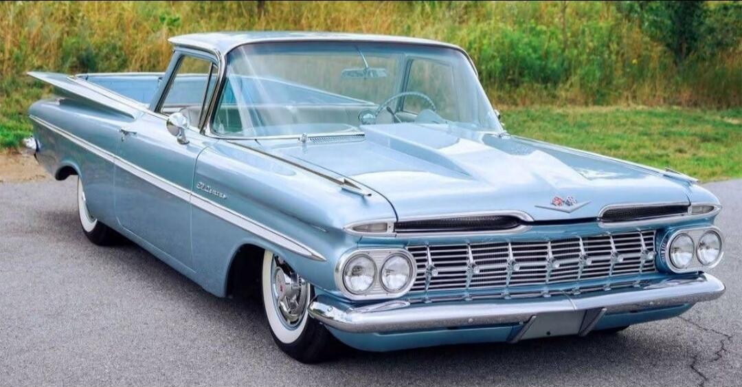A light blue vintage Chevrolet El Camino car-truck hybrid from the late 1950s, possibly a 1959 model, is parked on an asphalt surface with green foliage in the background. The vehicle features prominent tail fins, chrome accents, and whitewall tires.