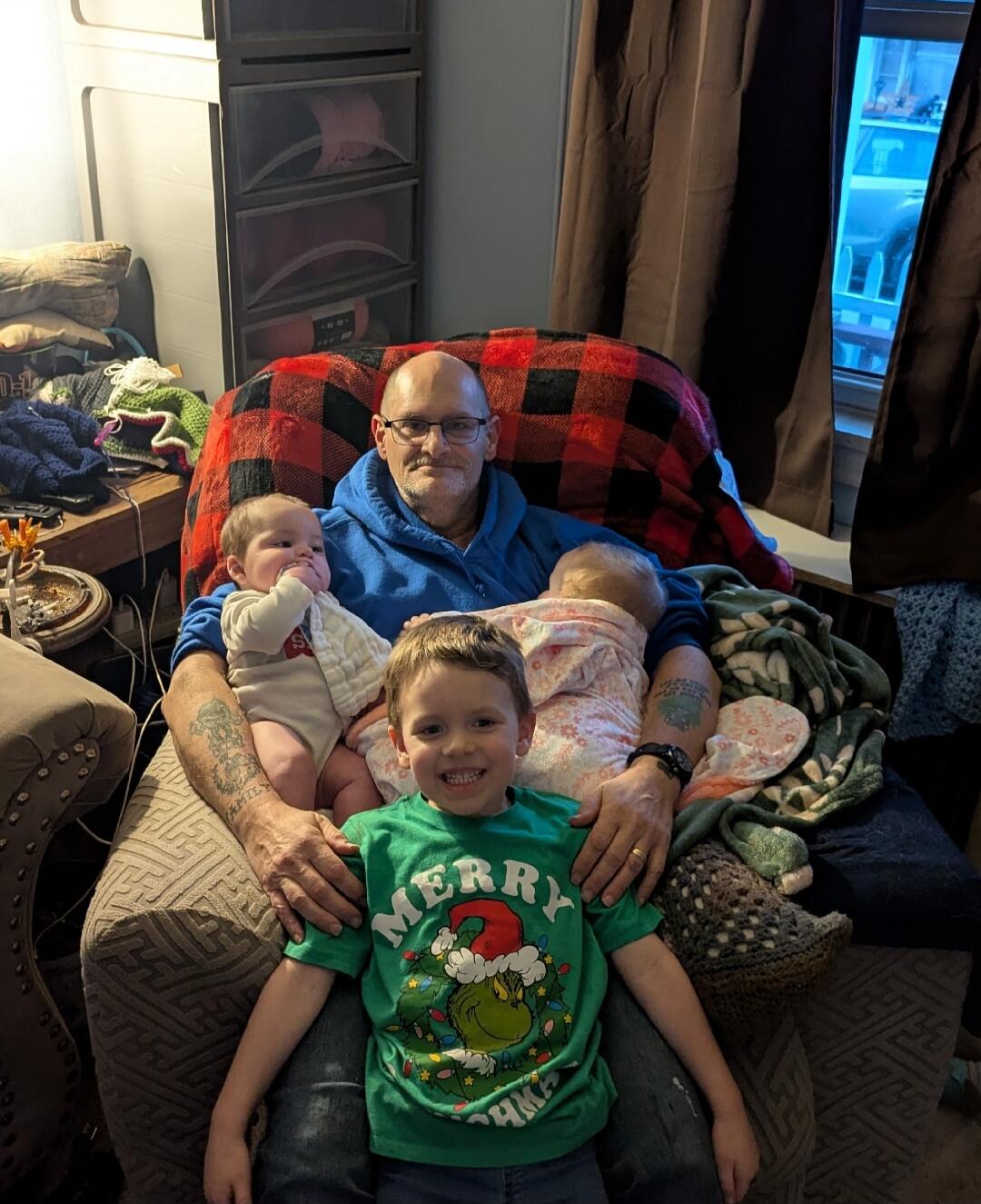 A man sits in a chair with two babies on his lap and a smiling young boy standing in front. The man is wearing a blue shirt, and the trio is gathered on a chair covered with a red-and-black plaid blanket, indoors.
