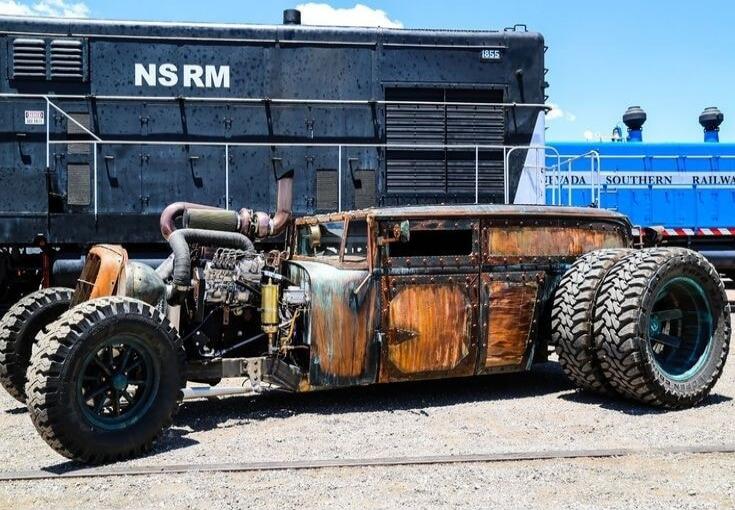 A heavily customized rat rod car with large off-road tires and an exposed engine is parked on gravel next to a black train locomotive marked 'NSRM 1855'. In the background, a blue train car with 'NEVADA SOUTHERN RAILWAY' written on it is visible under a clear blue sky.