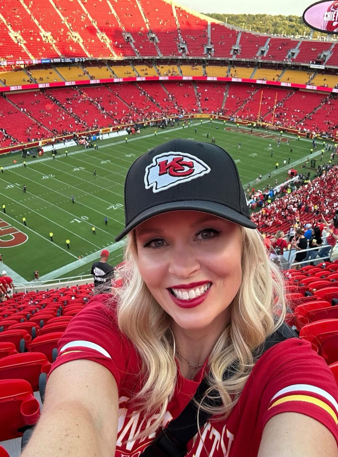 A Kansas City Chiefs fan poses for a selfie at Arrowhead Stadium. She wears a Chiefs cap with the team's logo and a red jersey, with the football field and crowd in the background.