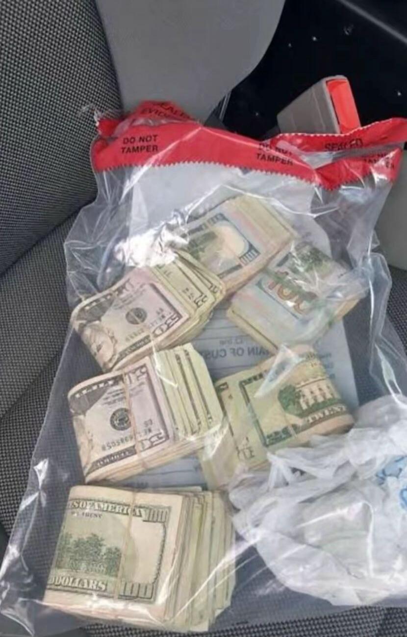 Stacks of US dollar bills, including 20s, 50s, and 100s, are visible inside a clear plastic bag. The bag has a red strip at the top with the text 'DO NOT TAMPER' and 'EVIDENCE'.