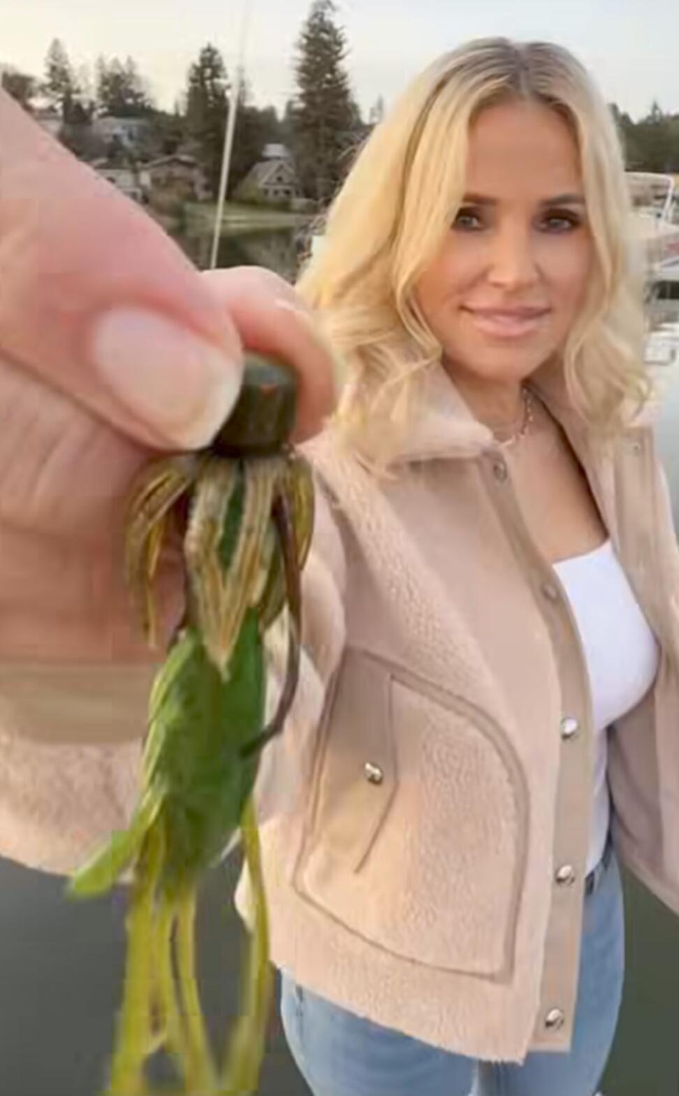 A woman holds up a green and brown fishing lure, smiling at the camera. She is wearing a light-colored jacket and a white top, standing outdoors near a body of water with houses and trees in the background.