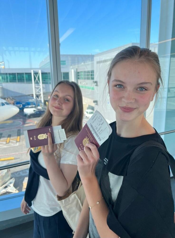 Two young women are smiling and holding passports and boarding passes in an airport terminal, with an airplane visible outside the window.