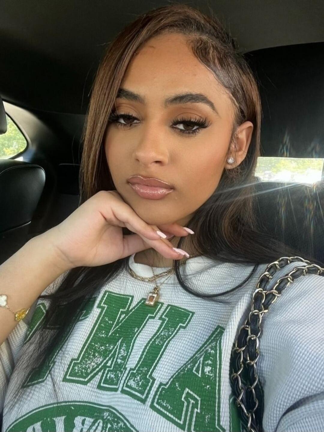 A young woman with long brown hair and makeup, wearing a light-colored waffle-knit top with green letters, possibly 'MIA'. She is taking a selfie in a car, with her hand resting on her chin. She has a necklace, a bracelet, and a black chain strap visible.